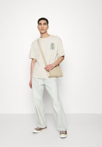 Beige oversized t-shirt with green graphic, light blue straight-leg jeans, and brown sneakers. Carrying a beige crossbody bag.