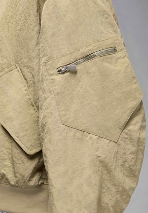 Khaki jacket with a textured finish, featuring a side zip pocket and a ribbed hem. The fabric appears lightweight and crinkled.