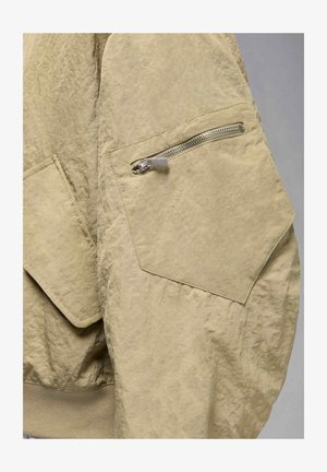 Khaki jacket with a textured finish, featuring a side zip pocket and a ribbed hem. The fabric appears lightweight and crinkled.