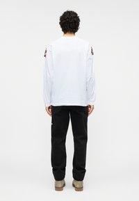 Λευκό μακρυμάνικο πουκάμισο με στρογγυλή λαιμόκοψη, με κόκκινες λεπτομέρειες σε ύφασμα στα μανίκια, συνδυασμένο με μαύρο παντελόνι και μπεζ παπούτσια.