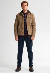Giacca in corduroy marrone con colletto peloso, camicia a quadri sotto, jeans blu scuri e stivali marroni con lacci. In piedi contro uno sfondo semplice.