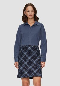Femme aux cheveux bruns mi-longs portant une chemise bleue à boutons et une jupe écossaise bleu marine, se tenant devant un fond clair uniforme.