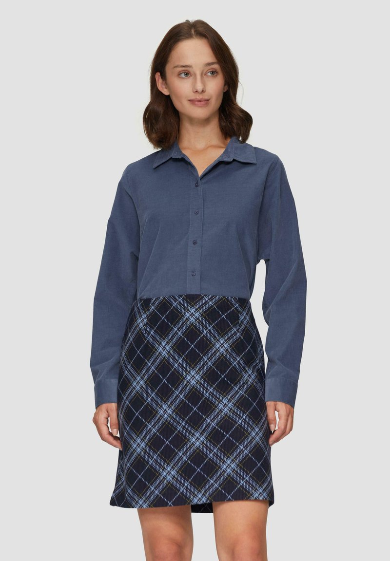 Femme aux cheveux bruns mi-longs portant une chemise bleue à boutons et une jupe écossaise bleu marine, se tenant devant un fond clair uniforme.