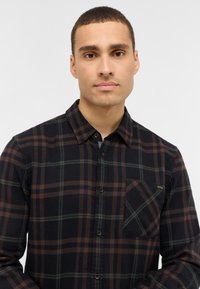 Chemise en flanelle à carreaux noirs et marron avec des accents verts, manches longues, fermeture boutonnée, et une poche poitrine avec une étiquette.