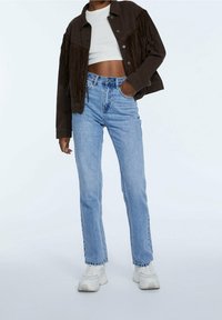 Chaqueta marrón con flecos, top corto blanco, vaqueros de tiro alto de color azul claro y zapatillas blancas gruesas. Atuendo casual que muestra estilos en capas.
