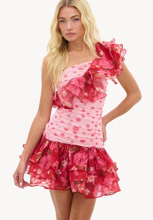 Femme blonde portant une robe rose asymétrique avec des volants floraux rouges et des pois, debout avec la main sur la hanche sur fond blanc.