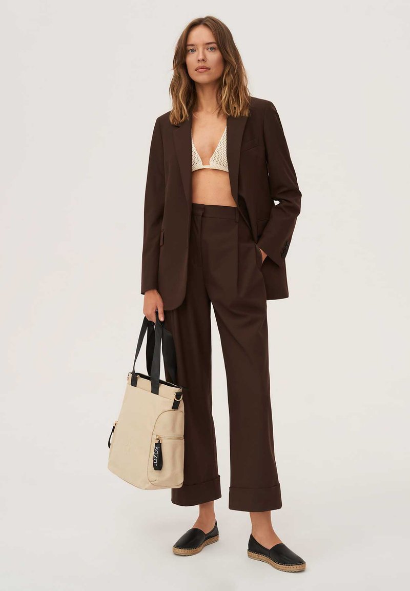 Kobieta w ciemnobrązowym garniturze z kremowym plecionym biustonoszem, trzymająca beżową torbę na ramię z czarnymi uchwytami i nosząca czarne buty wsuwane.