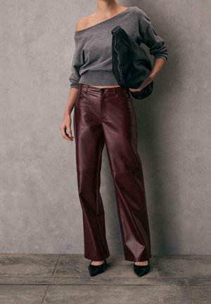 Personne portant un pull gris à épaules dénudées, pantalon en cuir bordeaux taille haute, talons noirs, tenant une grande pochette noire.