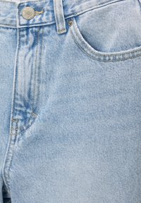 Gros plan sur un jean en denim bleu clair montrant un bouton, des passants de ceinture, une braguette zippée et une poche avant avec des coutures détaillées.