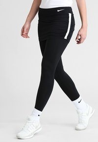 Czarna tenisówka z marszczonym pasem, zestawiona z czarnymi legginsami i białymi trampkami. Zawiera białe logo Nike i paski.