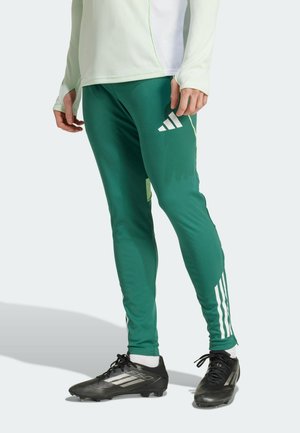 Persoon draagt groene Adidas trainingsbroek met witte strepen en logo, zwarte voetbalschoenen en een wit shirt met lange mouwen en staat rechtop.