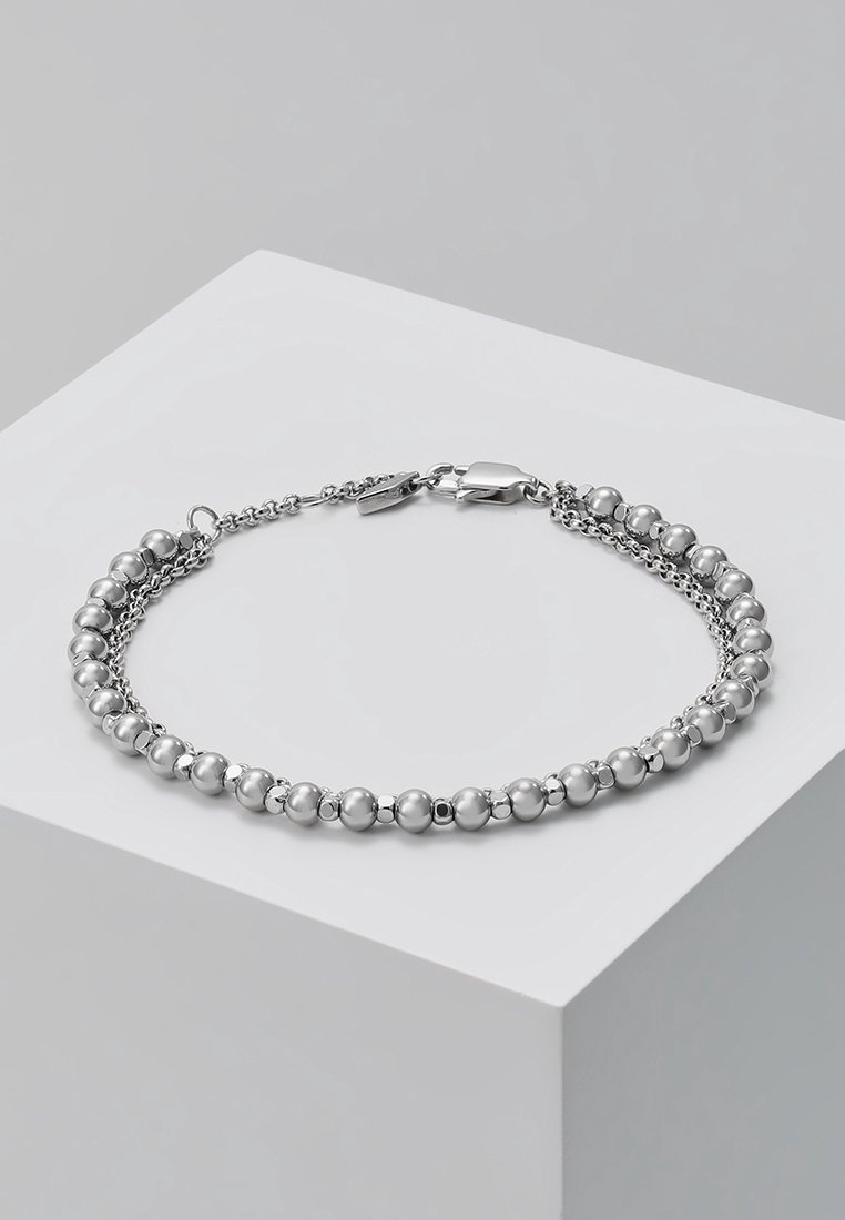 Bracelet ton argent avec deux chaînes parallèles, dont une avec des perles rondes, et une fermeture à mousqueton, présenté sur un socle blanc.