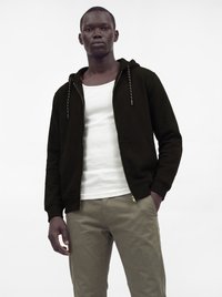 Homme portant un sweat à capuche noir zippé par-dessus un débardeur blanc et un pantalon beige, debout devant un fond blanc uni.