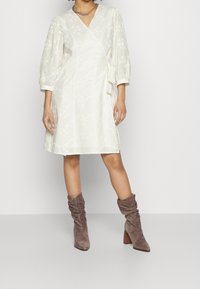 Krémové šaty s květinovou texturou, balónovými rukávy a bočním zavazováním. Nošeny s volnými taupe botami a stříbrným řetízkem.