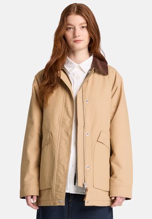 Jonge vrouw met lang rood haar draagt een beige jas met een donkere kraag over een wit overhemd met knopen en een donkere broek.