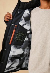 Die Innenseite der schwarzen Puffy-Jacke verfügt über ein grau-schwarzes Spritzmuster, eine orangefarbene Reißverschlusstasche und ein Pflegeetikett mit wasserdichten Details.