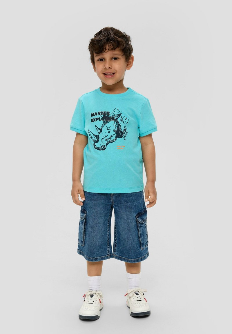 Türkises Baumwoll-T-Shirt mit einem schwarzen Rhinozeros-Graphic, kombiniert mit Jeans-Cargo-Shorts und weißen Sneakers mit schwarzen Akzenten.