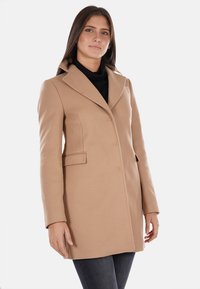 Cappotto beige su misura con colletto a soffietto, due tasche frontali e chiusura a bottoni. Realizzato in tessuto liscio, progettato per una silhouette aderente.
