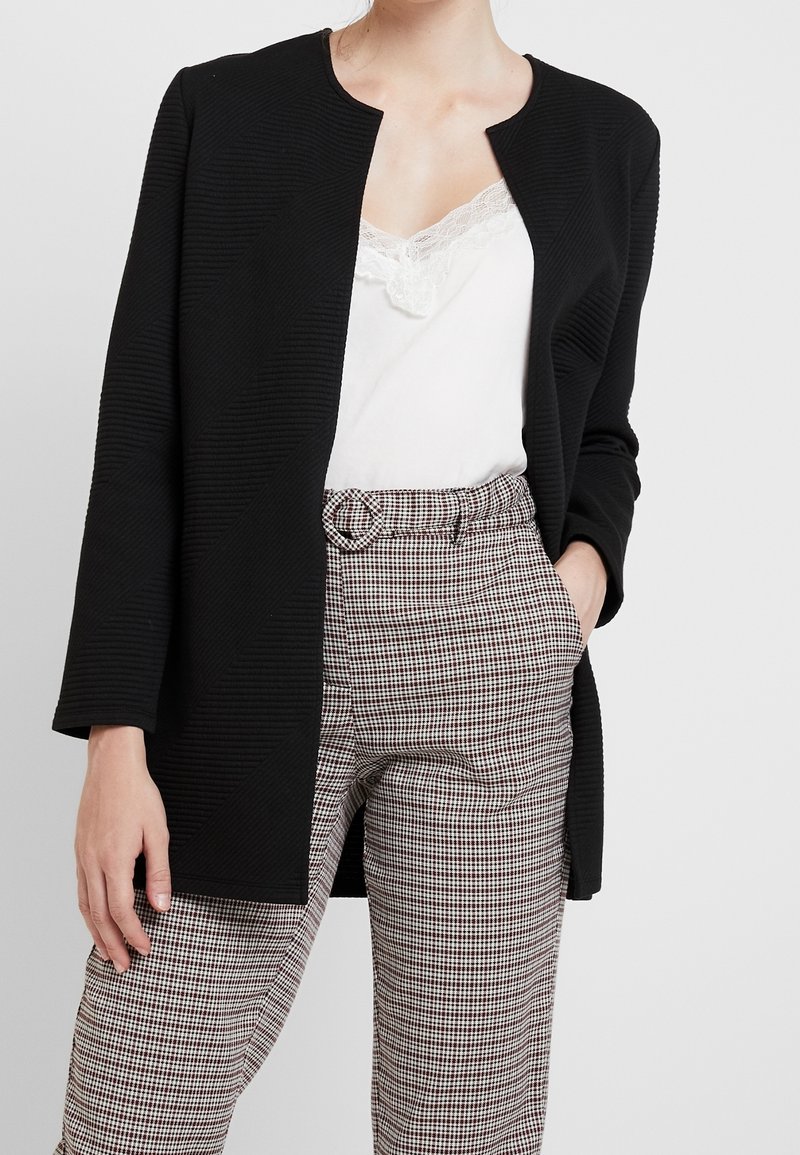 Femme portant une veste noire texturée ouverte à l'avant, un caraco blanc bordé de dentelle et un pantalon à carreaux taille haute avec ceinture, main dans la poche.