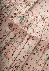 Primo piano di un tessuto beige chiaro con motivi floreali in verde, rosa e marrone, dotato di un bordo arricciato e bottoni di legno.