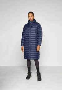 Tumesinine sulejope, millel on täislukuga esikülg, kapuuts ja horisontaalne tikandmustrit. Kantud mustade retuuside ja mustade saapadega.