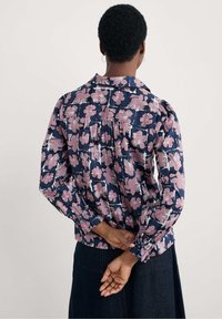 Persona con capelli corti e ricci, che indossa una blusa blu navy con stampa floreale rosa e una gonna scura, con le mani incrociate dietro la schiena.