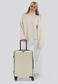 Vrouw in een crème coltrui en witte broek die staat met een metallic gouden harde koffer met vier wielen en een zwarte handgreep.