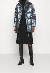 Vadderad, iriserande puffjacka i mörkblått med hög krage och dragkedja, bärs över en svart ribbad klänning och knähöga svarta stövlar.