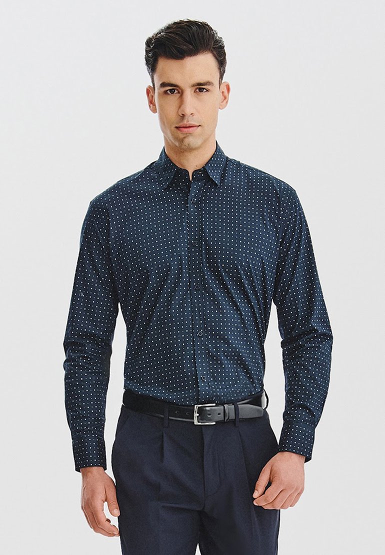 Granatowa koszula z długim rękawem w małe białe kropki, z kołnierzykiem zapinanym na guziki i slim fit. Noszona z czarnym paskiem i ciemnymi spodniami.