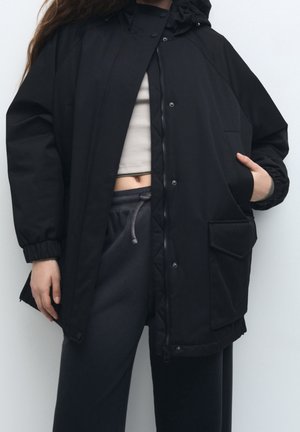 Osoba ubrana w czarną, oversize’ową kurtkę z kapturem, na beżowym topie odsłaniającym brzuch i ciemnoszare spodnie z sznurkiem, stojąca z rękami w kieszeniach.