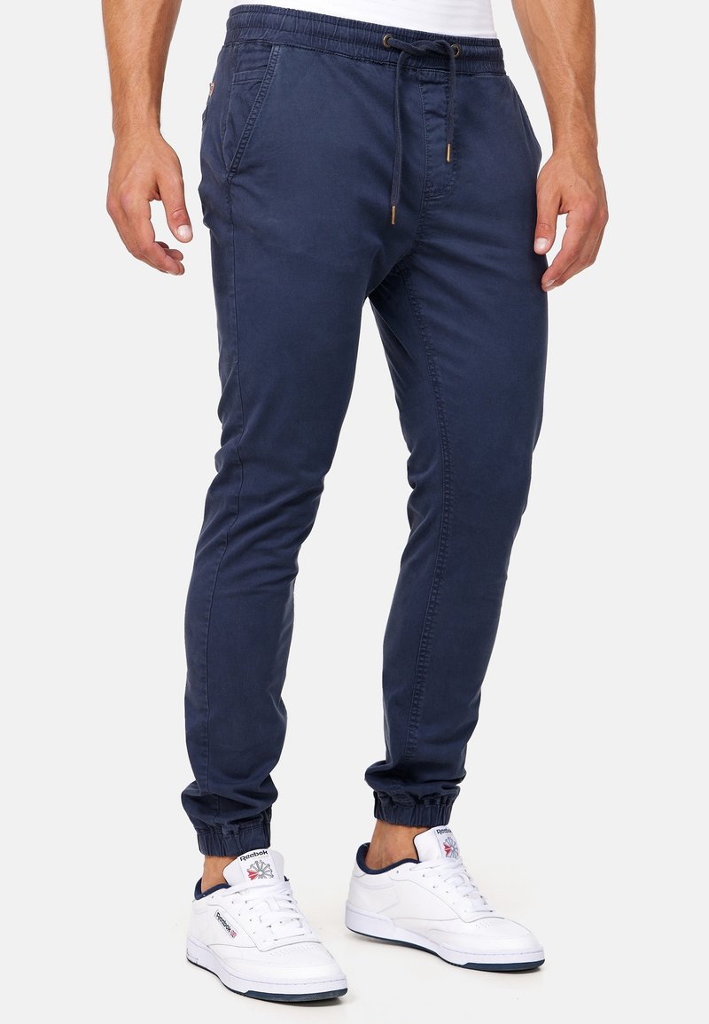 Homme portant un pantalon de jogging bleu marine avec taille à cordon et poignets élastiques, associé à des baskets blanches Reebok.