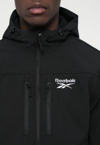 Czarna kurtka z kapturem z logo Reebok, z zamkiem z przodu i dwiema pionowymi kieszeniami na zamek na klatce piersiowej, noszona przez osobę.