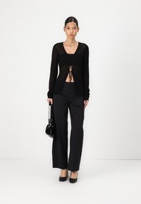 Maglione nero a maniche lunghe con apertura frontale, coulisse, texture a coste, abbinato a pantaloni neri a gamba larga e una borsa piccola.