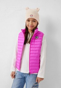 Roze donsvest met horizontaal doorgestikt patroon, voorzien van een rits aan de voorkant en een ronde logo-opdruk. Gecombineerd met een beige gebreide muts.