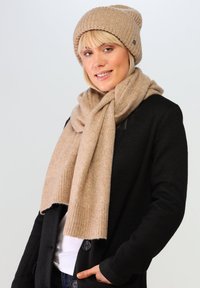 Beżowa dzianinowa czapka i pasujący szalik, wykonane z miękkiej wełny, noszone na czarnym, teksturowanym kardiganie. Szalik elegancko układa się wokół szyi.