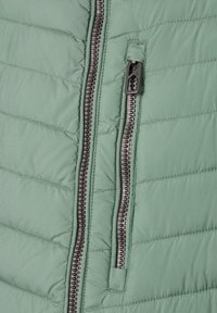 Nahaufnahme einer hellgrünen gesteppte Jacke mit einem großen zentralen Reißverschluss und einer kleineren vertikalen Reißverschlusstasche daneben.