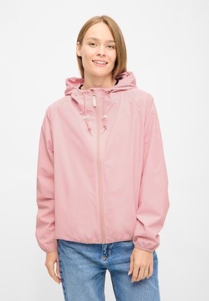 Smilende kvinde iført en lyserød hættejakke med snore og blå jeans mod en ensfarvet hvid baggrund.