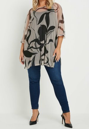 Kobieta ubrana w przezroczystą beżową bluzkę z dużymi czarnymi kwiatowymi wzorami, ciemnoniebieskie obcisłe jeansy i czarne wysokie obcasy, stojąca na białym tle.