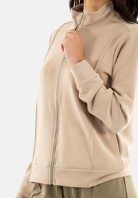 Chaqueta beis con cremallera, cuello alto, mangas largas y puños acanalados. Textura suave con un diseño ajustado, ideal para uso casual.