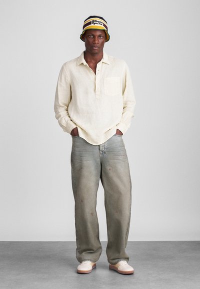 Camisa de linho bege de mangas longas com um bolso, combinada com jeans de perna larga desgastados em cinza claro e ténis brancos. Chapéu de balde multicolorido.
