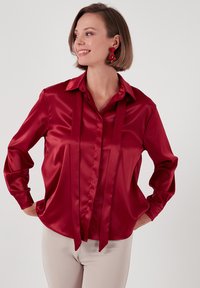 Red satin blouse with a collar, long sleeves, and two front ties. Smooth texture with a slightly loose fit, suitable for various occasions.