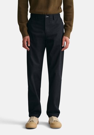 Black, tailored trousers with a smooth fabric, flat front, and subtle side pockets, paired with beige sneakers.