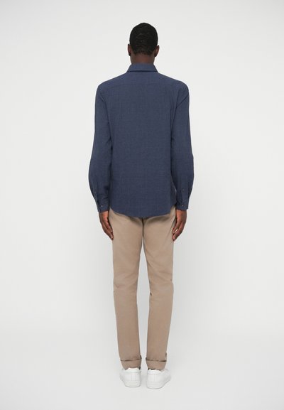 Camisa de mangas compridas azul-marinho com colarinho abotoado, textura suave, combinada com calças chinos de um bege claro e ténis brancos, vista pela parte de trás.