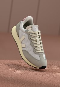 Sneaker con una toma superiore in mesh bianco, dettagli in suede grigio, lacci color crema e una suola gialla. Presenta un logo sulla linguetta e sul lato.