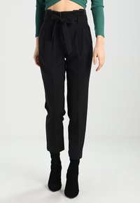 Donna che indossa pantaloni neri a vita alta con una cintura legata, abbinati a stivaletti neri e a un top verde a maniche lunghe e cropped.