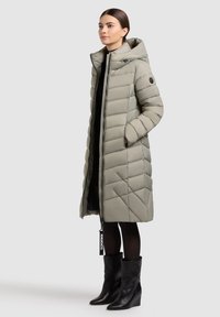 Dolga, svetlo siva puhovka s kapuco, z diagonalnim quilted dizajnom, sprednjim zadrgo in stranskimi žepi, v kombinaciji s črnimi čevlji z visokimi petami.