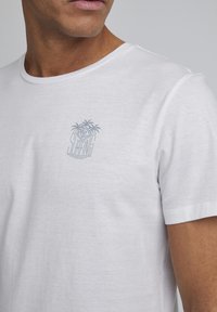 Hvit T-skjorte laget av mykt materiale, med et lyst grått palmetre og "Surfing" grafikk på brystet. Korte ermer og klassisk rund hals.