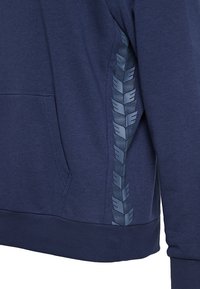 Felpa blu navy con tessuto testurizzato, tasca a marsupio e un motivo logo a contrasto lungo il lato. Polsini e orlo elasticizzati.