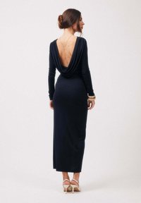 Femme aux cheveux foncés attachés en chignon, portant une longue robe noire dos nu, un collier en or, des bracelets et des chaussures blanches à talons hauts, debout de profil.