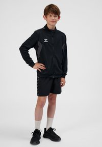 Svart zip-up träningsjacka med grå detaljer, kombinerad med svarta shorts med en zigzag-rand. Modellen bär ljusa strumpor och svarta sportskor.